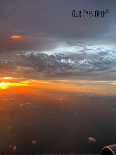 Clouds float by while the sun peaks out among them in hues of purple, blue, yellow and orange.