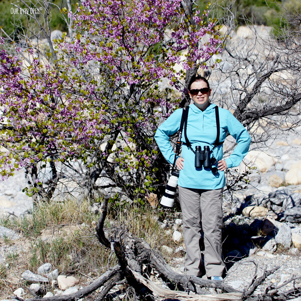 Lisa Coleman with binoculars strapped in & camera at the ready standing in front of a tree at Red Rock Canyon outside of Las Vegas, Nevada.