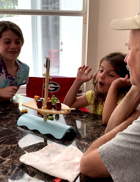 Two granddaughters playing a balance game.  It is a wooden ship with 25 pieces.  5 pieces of different cargo items are light and heavy weighted. Roll the dice to see what you have to mount on the boat without tipping it over.